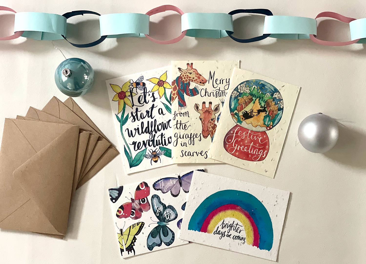 seasonal cards fanned out on a white background with brown envelopes beside them and a blue and pink paper chain above