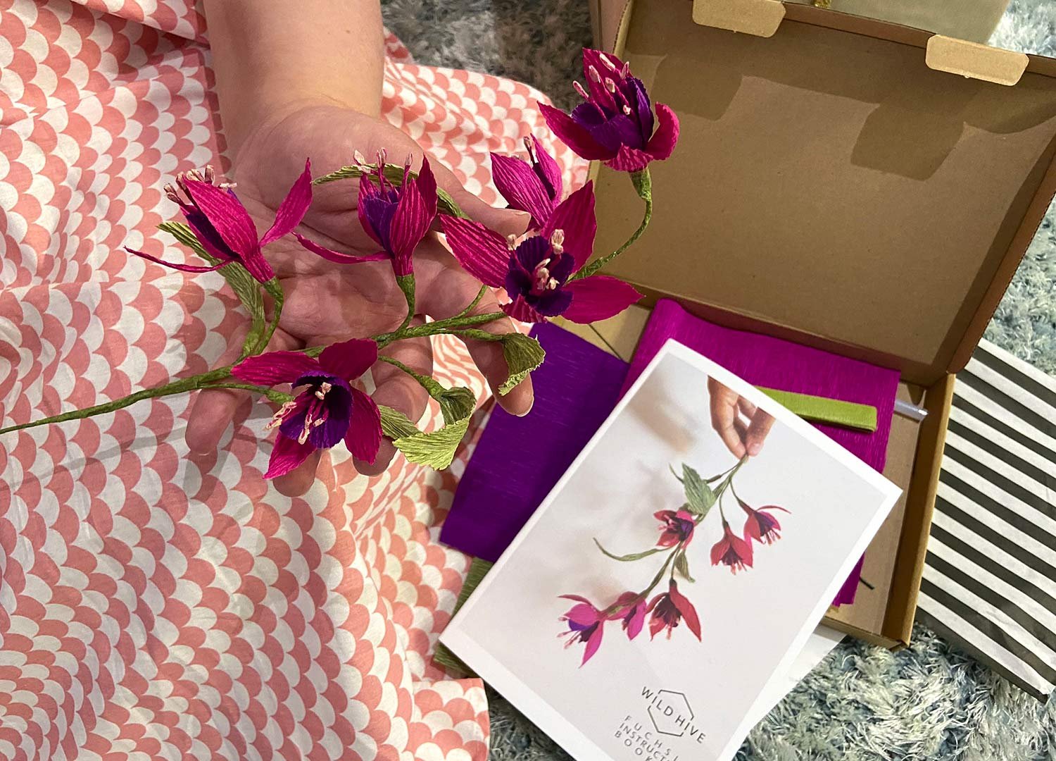 a model holds a paper fuchsia over her palm and below there is the instructions booklet and crafting materials