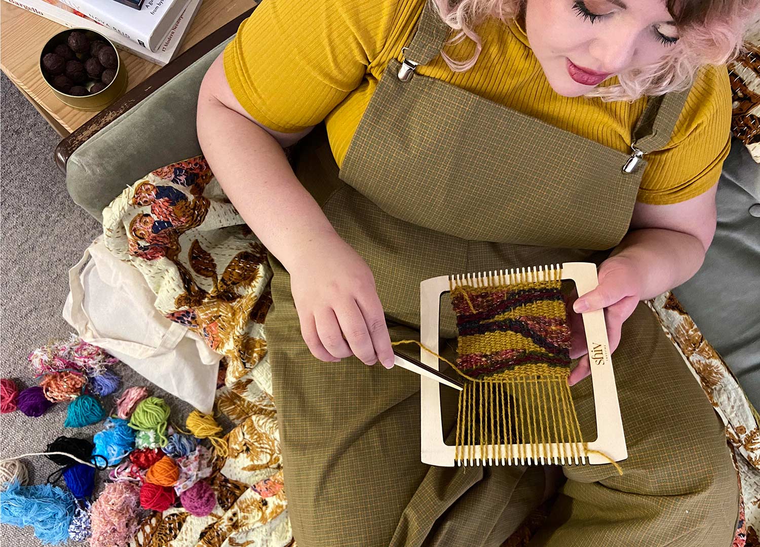 a model is holding a loom with a tapestry in progress with a selection of colourful yarn beside them.