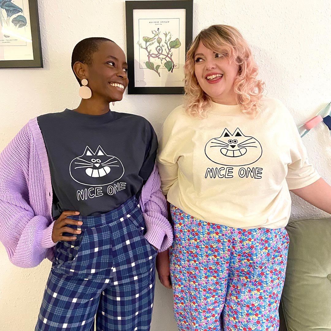 two models are looking at each other smiling, wearing t shirts that have a cat face on and the words nice one underneath